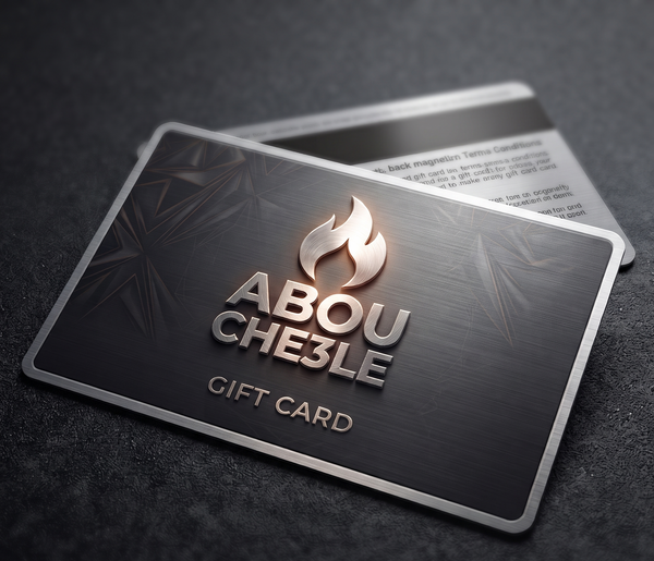 A metallic gift card with a brushed dark gray finish, featuring the brand name 'ABOU CHE3LE' in raised silver lettering beneath a stylized flame logo. The card is shown at an angle on a dark textured surface, with a second card partially visible behind it. -  Abou Che3le Gift Card -  Abouche3le