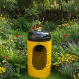 A yellow barrel-style outdoor pet stove or fire pit with a black lid, shown on a tiled floor in front of a shop window. Blue barrels and store fixtures are visible in the background.