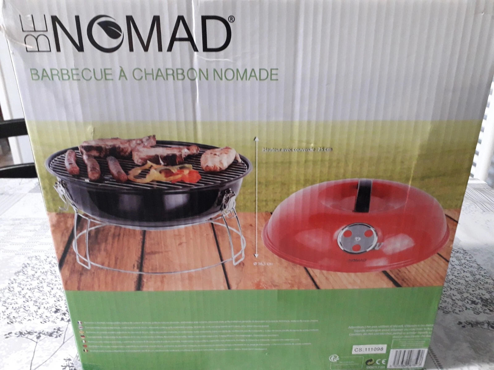 BE NOMAD charcoal barbecue box packaging showing a black round grill with food on the grate and a red lid on a wooden surface, photographed in a real indoor setting.