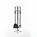 A black wrought-iron fireplace tool set with wooden handles and a light tan brush bristle head, displayed on a stand in front of a large concrete pillar.