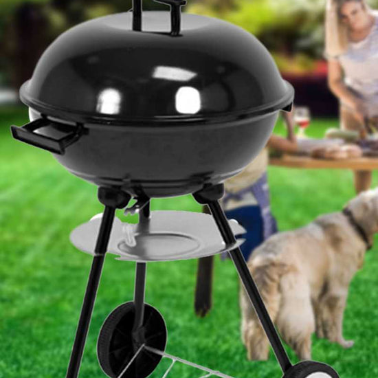A black kettle-style outdoor charcoal LUND CHARCOAL BBQ GRILL WITH LID 41CM, three legs, and two wheels, shown on a green lawn with a blurred person and dog in the background.