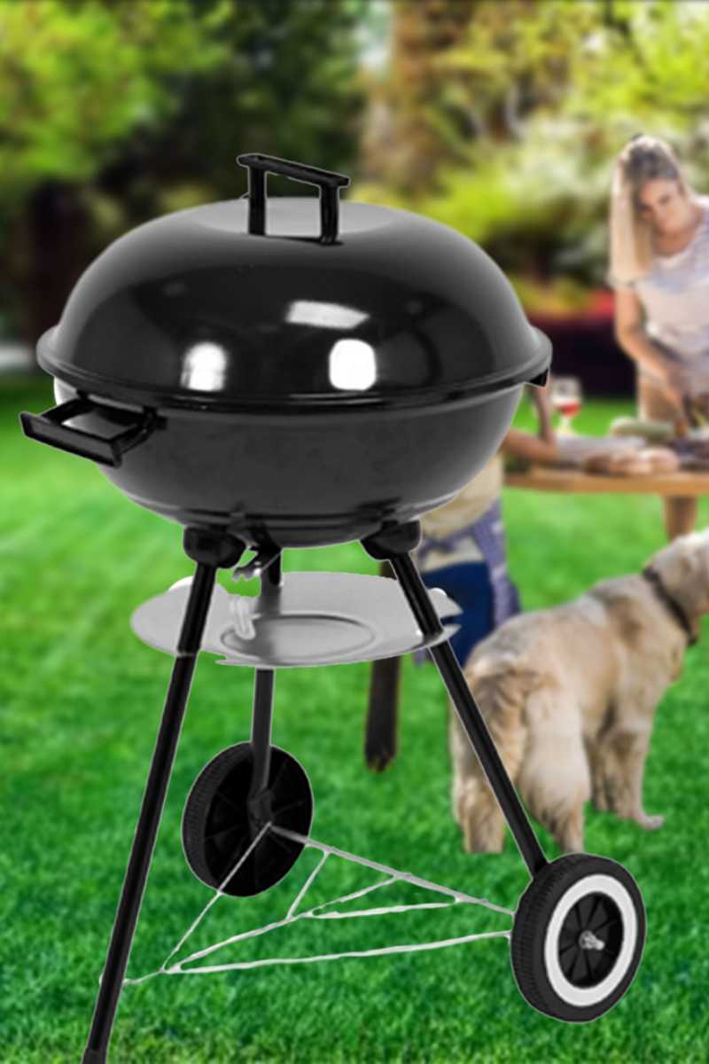 A black kettle-style outdoor charcoal LUND CHARCOAL BBQ GRILL WITH LID 41CM, three legs, and two wheels, shown on a green lawn with a blurred person and dog in the background.