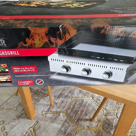 A boxed gas grill product labeled 'Grill Meister' and 'Plancha-GasGrill' sits on a wooden table in a home setting, with the retail box showing a photo of the grill and flame graphics.