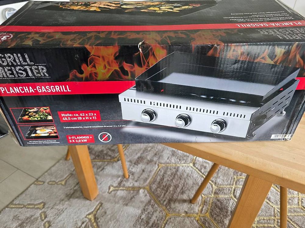 A boxed gas grill product labeled 'Grill Meister' and 'Plancha-GasGrill' sits on a wooden table in a home setting, with the retail box showing a photo of the grill and flame graphics.